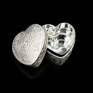 May include: A silver heart-shaped trinket box with an open lid. The lid features an embossed floral design. The box is on a black background. The box is likely made of metal.