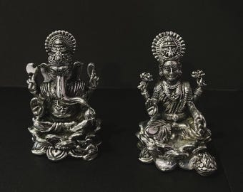 Op de afbeelding: Twee zilverkleurige beelden van hindoeïstische goden. Het beeld links toont Ganesha, en het beeld rechts Lakshmi. Beide beelden zijn gedetailleerd en staan op een donkere ondergrond.