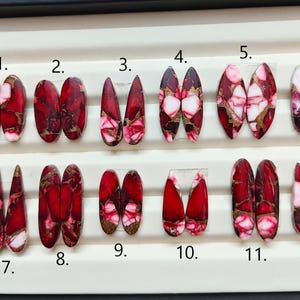 Può includere: Dodici paia di cabochon lucidati, di forma ovale e a goccia, disposti su due file su un vassoio bianco. Ogni pietra presenta un vivace motivo marmorizzato rosso e bianco con venature color rame metallizzato. Ogni coppia è numerata da 1 a 12.