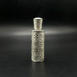 May include: A silver perfume bottle with a detailed floral pattern. The bottle has a cylindrical body and a separate, decorative cap. The design is symmetrical, with the floral pattern repeated around the bottle's circumference. The bottle is made of metal and has a shiny finish.