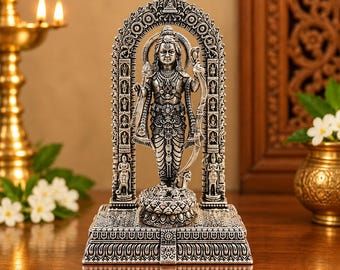 May include: A silver-toned metallic figurine of a standing deity holding a spear and a bow, positioned within an ornate arched frame. The figure stands on a lotus-shaped base atop a rectangular pedestal, featuring intricate carvings and detailed relief work.