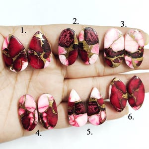 May include: A collection of six pairs of polished gemstone cabochons in various shapes. The stones feature a mix of red, pink, gold, and brown colors. The cabochons are displayed against a neutral background, showcasing their unique patterns and textures.