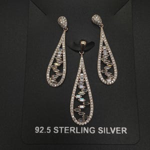May include: A jewelry set featuring a pendant and matching earrings. The teardrop-shaped pieces are outlined with small, clear stones and filled with a zig-zag pattern of rectangular stones. The text "92.5 STERLING SILVER" is printed below the jewelry.