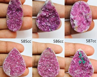 May include: Six individual cabochons featuring vibrant magenta druzy crystal surfaces. The stones are labeled 582cc, 583cc, 854cc, 585cc, 586cc, and 587cc. Each piece displays a textured, sparkling crystalline face with varied shapes including oval, pear, and rounded forms.