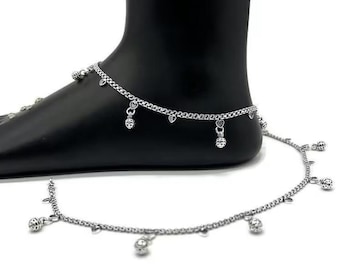 May include: Silver anklet with dangling heart and ball charms. The anklet is displayed on a black foot form. The chain is delicate and features small heart and ball pendants. A second anklet lies below.