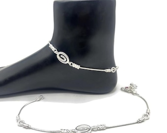 May include: A silver anklet with a spiral design is displayed on a black foot form. The anklet features a delicate chain with decorative elements and a spiral centerpiece. A second anklet with a similar design lies below.