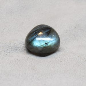 以下が含まれることがあります： 研磨された、丸みを帯びたラブラドライトの宝石。石は濃いグレーを基調とし、青とティールの虹色の輝きがあります。表面は滑らかで反射性があり、微妙な光沢があります。宝石は自然で有機的な形をしています。