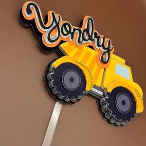 May include: A yellow dumper truck cake topper with black wheels and a black and orange "Yondry" name on top. The topper is on a clear plastic stick.