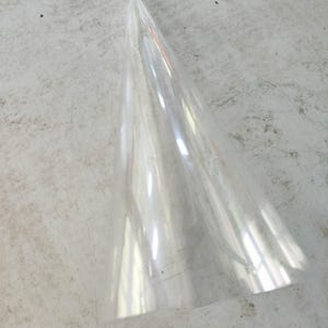Puede incluir: Un objeto de plástico transparente en forma de cono. El objeto es transparente y parece estar hecho de un material fino y flexible. El cono se estrecha hasta un punto afilado y tiene una superficie lisa. El objeto está sobre una superficie gris claro.