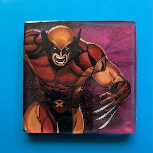 May include: A square resin coaster featuring a comic book illustration of Wolverine, a superhero with sharp claws, in a red and yellow costume with a purple background.