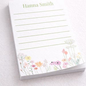 Può includere: Un blocco note bianco con pagine a righe verdi e un design floreale ad acquerello. Il blocco note ha il nome "Hanna Smith" stampato in verde in alto. Il design floreale presenta fiori rosa, gialli e arancioni.