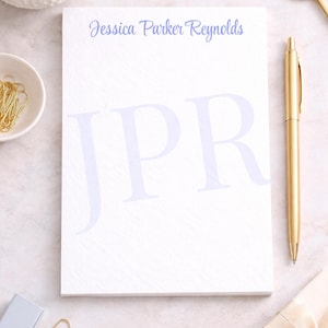 May include: A white notepad with the name "Jessica Parker Reynolds" in blue script and the initials "JPR" in a larger, lighter blue font. A gold pen and other stationery items are arranged around the notepad.