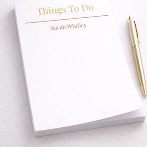 Può includere: Un blocco note bianco con la scritta dorata "Things To Do" e una linea rossa. Il blocco note ha il nome "Sarah Whitley" stampato sotto la linea. Una penna dorata è appoggiata sul lato destro del blocco.