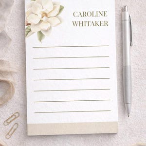 Può includere: Un blocco note bianco con pagine a righe, un'illustrazione di un fiore di magnolia e il nome "CAROLINE WHITAKER" in verde. Sono presenti anche una penna d'argento e due fermagli dorati. Il blocco note ha un bordo inferiore beige.
