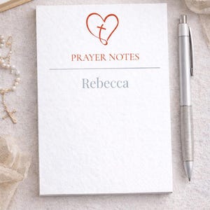 Può includere: Un blocco note bianco con le parole "PRAYER NOTES" e il nome "Rebecca" stampati sopra. Un cuore rosso con una croce è sopra il testo. Una penna d'argento è appoggiata sul lato destro del blocco note.