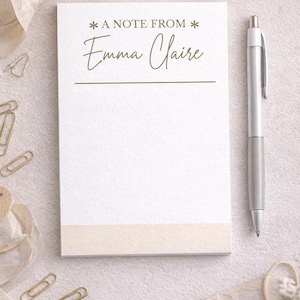 May include: A white notepad with the text "A NOTE FROM Emma Claire" in green script. A silver pen rests on the right side of the notepad. Gold paper clips and decorative ribbon are scattered around the notepad.