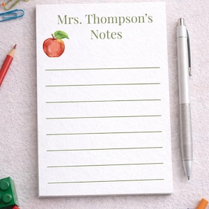 Può includere: Un blocco note bianco con il testo "Note della signora Thompson" e un'illustrazione ad acquerello di una mela. Una penna d'argento è appoggiata accanto al blocco note, con matite colorate, pennarelli e graffette sparsi. Il blocco note ha linee orizzontali.