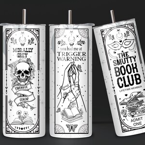 May include: Three white tumblers with black designs. The first tumbler has a skull and crossbones with the text "Morally Grey" and "Favorite Color". The second tumbler has two hands holding a book with the text "You had me at Trigger Warning". The third tumbler has a mask and the text "The Smutty Book Club, Lifetime Member, Admit One".