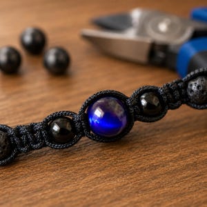May include: Close-up of a black macrame bracelet with a central blue gemstone and black beads. The bracelet is on a wooden surface, with additional black beads and tools in the background.