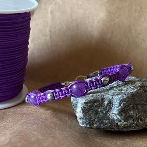 Peut inclure: Un bracelet macramé fait à la main de couleur violette, orné de trois grosses perles rondes violettes et de petites perles argentées. Le bracelet est présenté sur une roche grise avec une bobine de cordon violet en arrière-plan.