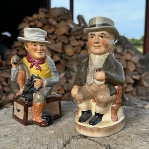 May include: Two ceramic figurines of men wearing hats and traditional clothing. The figurine on the left is holding a shoe and sitting on a wooden box. The figurine on the right is holding a pipe and sitting on a chair. Both figurines are painted in realistic detail.