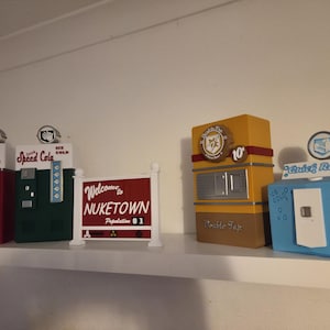 Peut inclure: Une collection de distributeurs automatiques originaux et un panneau sur une étagère blanche. Les machines sont rouges, vertes, jaunes et bleues, avec divers logos et textes, dont "Welcome to Nuketown" et "Double Tap".