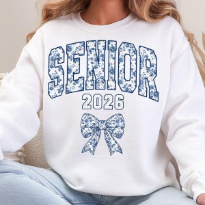 Op de afbeelding: Witte sweatshirt met het woord "SENIOR" in grote, blauwe bloemenletters, het jaartal "2026" eronder en een bijpassende strik. Een casual stijl, perfect voor een afstudeerfeest.