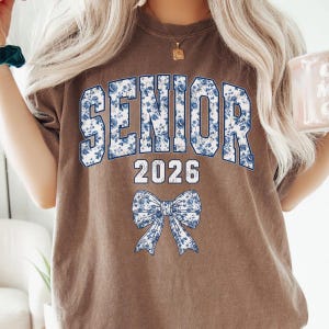 Op de afbeelding: Een bruin t-shirt met het woord "SENIOR" in grote, blauwe bloemenletters. Onder het woord "SENIOR" staat het nummer "2026" in het wit. Onderaan het shirt staat een blauwe bloemenstrik.