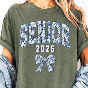 Op de afbeelding: Olijfgroen T-shirt met het woord "SENIOR" in blauwe bloemenprint, het jaartal "2026" en een blauwe bloemenstrik. Dit casual T-shirt is ideaal voor afstudeerfeesten.