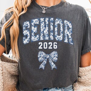 Op de afbeelding: Donkergrijs t-shirt met het woord "SENIOR" in blauwe bloemenprint, het jaartal "2026" in wit en een bijpassende strik. Een vintage-stijl shirt, perfect voor een afstudeerfeest.
