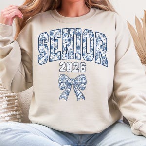 Op de afbeelding: Een beige sweatshirt met het woord "SENIOR" in grote, blauwe bloemenletters, het jaartal "2026" eronder en een blauwe bloemenstrik. De sweatshirt heeft een casual stijl.