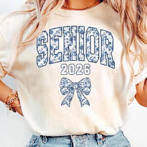 Op de afbeelding: Cr&egrave;mekleurig T-shirt met het woord "SENIOR" in blauwe bloemenprint, het jaar "2026" en een bijpassende strik-afbeelding. Het shirt is een casual stijl, geschikt voor afstudeerfeesten.