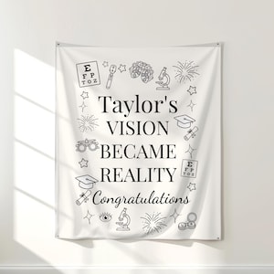 May include: A white banner featuring black text and illustrations. The banner states "Taylor's VISION BECAME REALITY Congratulations." Illustrations include an eye chart, microscope, mortarboard, and fireworks. The banner is displayed on a white wall.