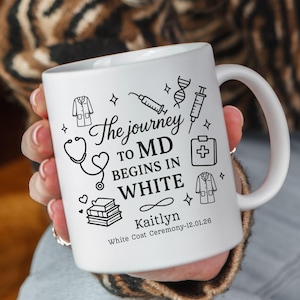 Puede incluir: Taza de cerámica blanca con dibujos en negro de símbolos médicos y texto. El texto dice "The journey to MD begins in white" con el nombre "Kaitlyn" y "White Coat Ceremony-12.01.26".