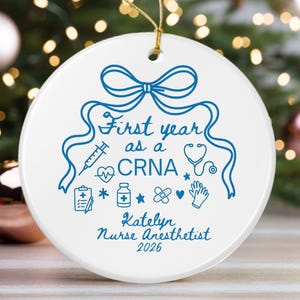 Puede incluir: Adorno navideño de cerámica blanca con un lazo azul y el texto "First year as a CRNA." Incluye ilustraciones médicas y el nombre "Katelyn Nurse Anesthetist 2026." Una cuerda dorada está adjunta.