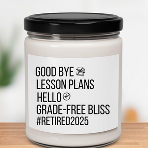 Puede incluir: Una vela blanca en un tarro de cristal con tapa negra. La etiqueta dice "GOOD BYE LESSON PLANS HELLO GRADE-FREE BLISS #RETIRED2025" en texto negro. La vela está sobre una superficie de madera.