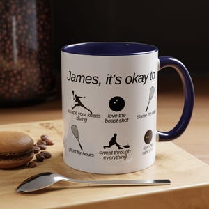Op de afbeelding: Een witte keramische mok met een blauwe rand en handvat. De mok heeft een zwart-witte illustratie van een persoon die squash speelt met de tekst "James, it's okay to" en een lijst met humoristische squash-gerelateerde zinnen.
