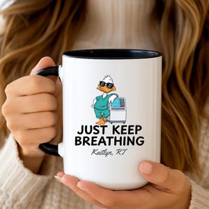 May include: White ceramic mug with a black interior and handle. The mug features a cartoon duck wearing sunglasses and scrubs, standing next to a medical machine. Text on the mug reads "JUST KEEP BREATHING" and "Kaitlyn, RT".