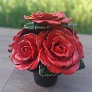 May include: A ceramic floral arrangement featuring three large, glossy red roses and several green leaves. The roses are arranged in a black, cylindrical vase. The background is blurred, with a wooden surface.