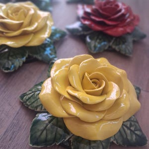 Può includere: Primo piano di sculture di rose in ceramica gialle e rosse. Ogni rosa ha petali dettagliati ed è posta su una base di foglie verdi. Le rose sono disposte su una superficie di legno, mettendo in risalto i loro colori vivaci.