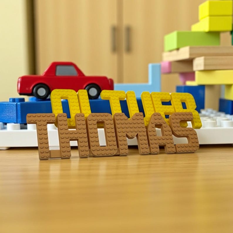 May include: A wooden toy scene featuring the name "THOMAS" in brown block letters, with "OLIVER" in yellow blocks behind it. A red toy car sits atop a blue block structure, with a colorful block tower in the background.
