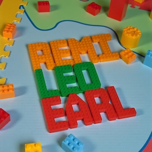 May include: A colorful play mat with the names "REMI", "LEO", and "EARL" spelled out in orange, green, and red building blocks. Various colored blocks are scattered around the names on a light blue surface, with a map-like design.