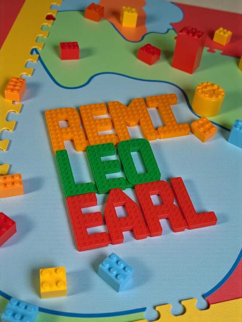 May include: A colorful play mat with the names "REMI", "LEO", and "EARL" spelled out in orange, green, and red building blocks. Various colored blocks are scattered around the names on a light blue surface, with a map-like design.