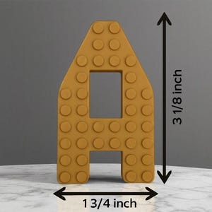 May include: Mustard yellow letter "A" made to resemble a building block, with a rectangular cutout in the center. The letter is 3 1/8 inches tall and 1 3/4 inches wide.