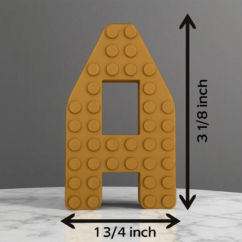 May include: Mustard yellow letter "A" made to resemble a building block, with a rectangular cutout in the center. The letter is 3 1/8 inches tall and 1 3/4 inches wide.