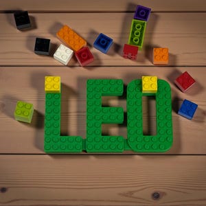May include: A wooden surface displays the word "LEO" spelled out in green building blocks, with yellow blocks on top of the letters. Scattered around are various colorful blocks in orange, red, blue, white, and black.