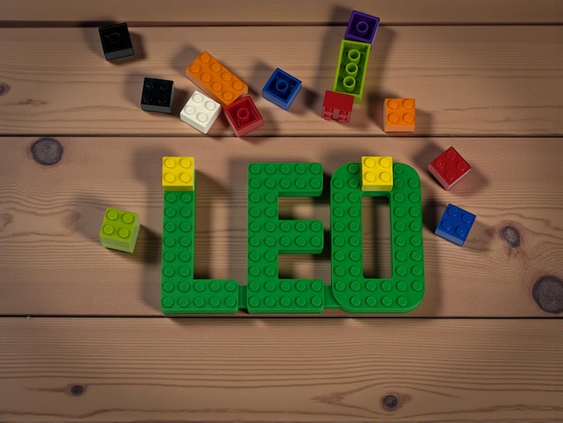 May include: A wooden surface displays the word "LEO" spelled out in green building blocks, with yellow blocks on top of the letters. Scattered around are various colorful blocks in orange, red, blue, white, and black.