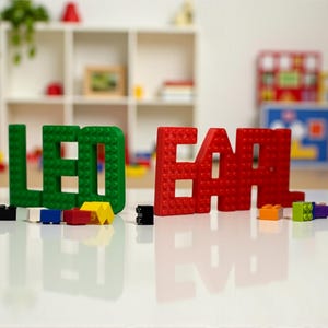 May include: Green and red building block letters spell out the word "LEGO" and "EAR". Various colorful building blocks are scattered on a white surface. The background includes a white shelf with toys.