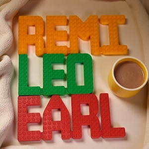 May include: Orange, green, and red block letters spell out the names REMI, LEO, and EARL. A small yellow cup filled with a brown liquid sits next to the letters. The letters and cup are on a white surface.