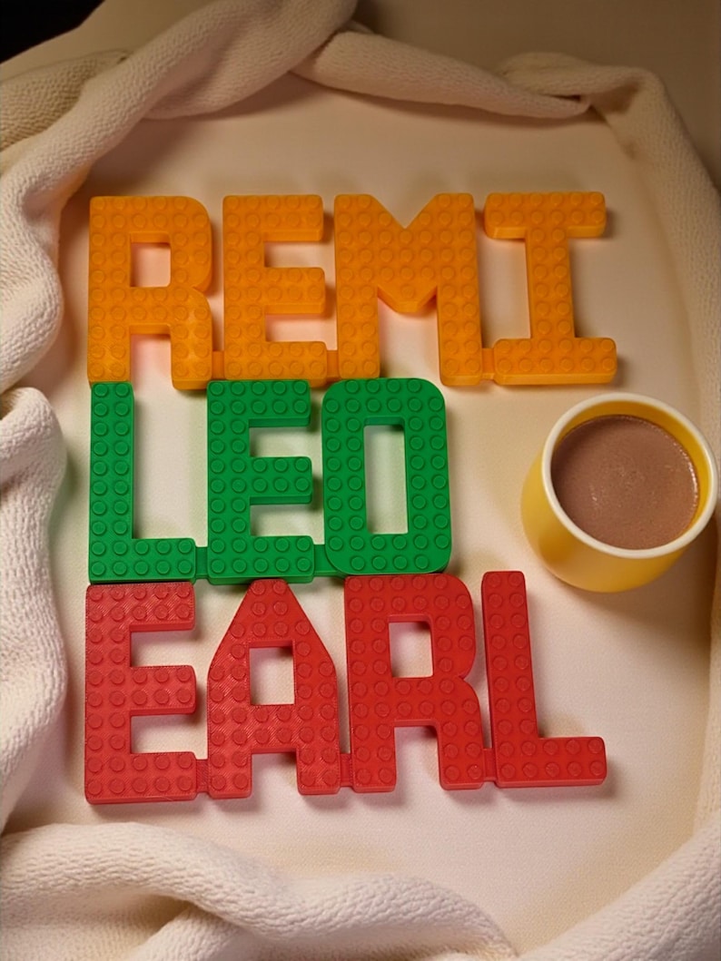 May include: Orange, green, and red block letters spell out the names REMI, LEO, and EARL. A small yellow cup filled with a brown liquid sits next to the letters. The letters and cup are on a white surface.